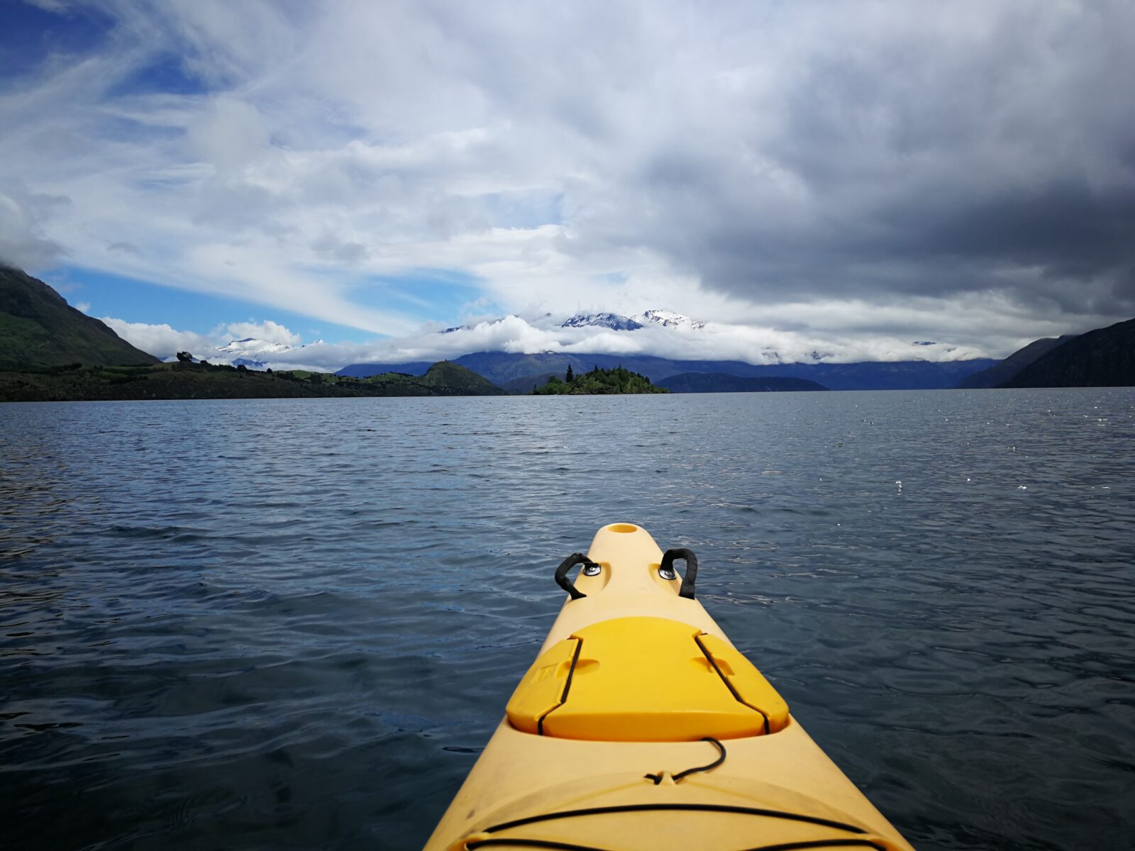 Die gelbe Spitze eines Kajaks ragt im Vordergrund auf dem Lake Wānaka ins Bild. Im Hintergrund erstreckt sich das ruhige Wasser mit Mou Waho Island und den schneebedeckten Gipfeln der Südalpen nahe dem Mount Aspiring National Park in Neuseeland.