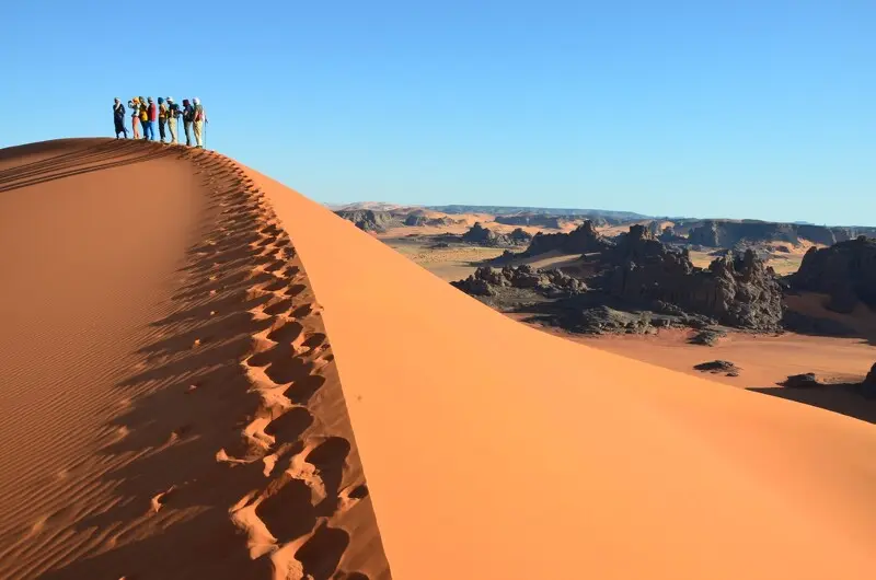 Eine Gruppe von neun Wanderern steht auf einem scharfen Dünengrat in der Tadrart Rouge im Tassili n’Ajjer Nationalpark in Algerien. Der Dünengrat mit menschlichen Fußspuren und Tierspuren verläuft diagonal im Vordergrund. Im Hintergrund erstreckt sich die Sahara-Wüstenlandschaft nahe Djanet mit dunklen Felsformationen unter blauen Himmel.