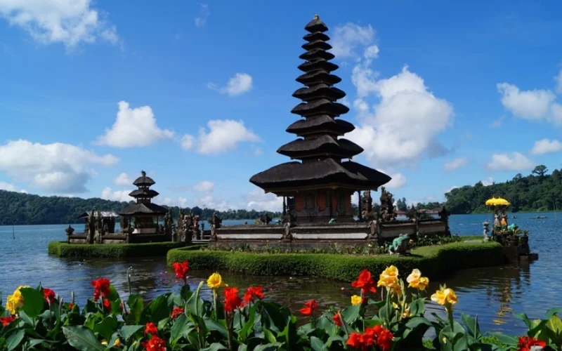 Mehrstufiger balinesischer Tempelturm (Meru) des Pura Ulun Danu Bratan direkt am Ufer eines Kratersees auf Bali. Im Vordergrund blühen rote und gelbe Canna-Blumen vor strahlend blauem Himmel mit leichten Wolken.