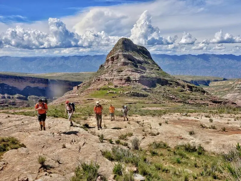 Eine Gruppe von Wanderern mit Rucksäcken zieht über eine felsige Hochebene im Torotoro-Nationalpark in Bolivien. Im Hintergrund erhebt sich eine gewaltige, pyramidenförmige Sandsteinformation mit deutlichen Gesteinsschichten unter einem leicht bewölkten blauen Himmel.