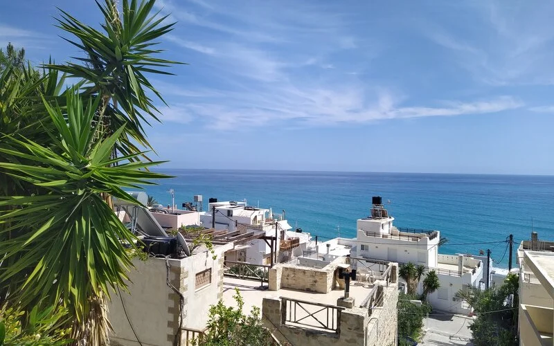 Blick über die hellen Flachdächer und Terrassen des Küstendorfs Myrtos auf Kreta zum tiefblauen Libyschen Meer, eingerahmt von einer Yucca-Palme im Vordergrund.