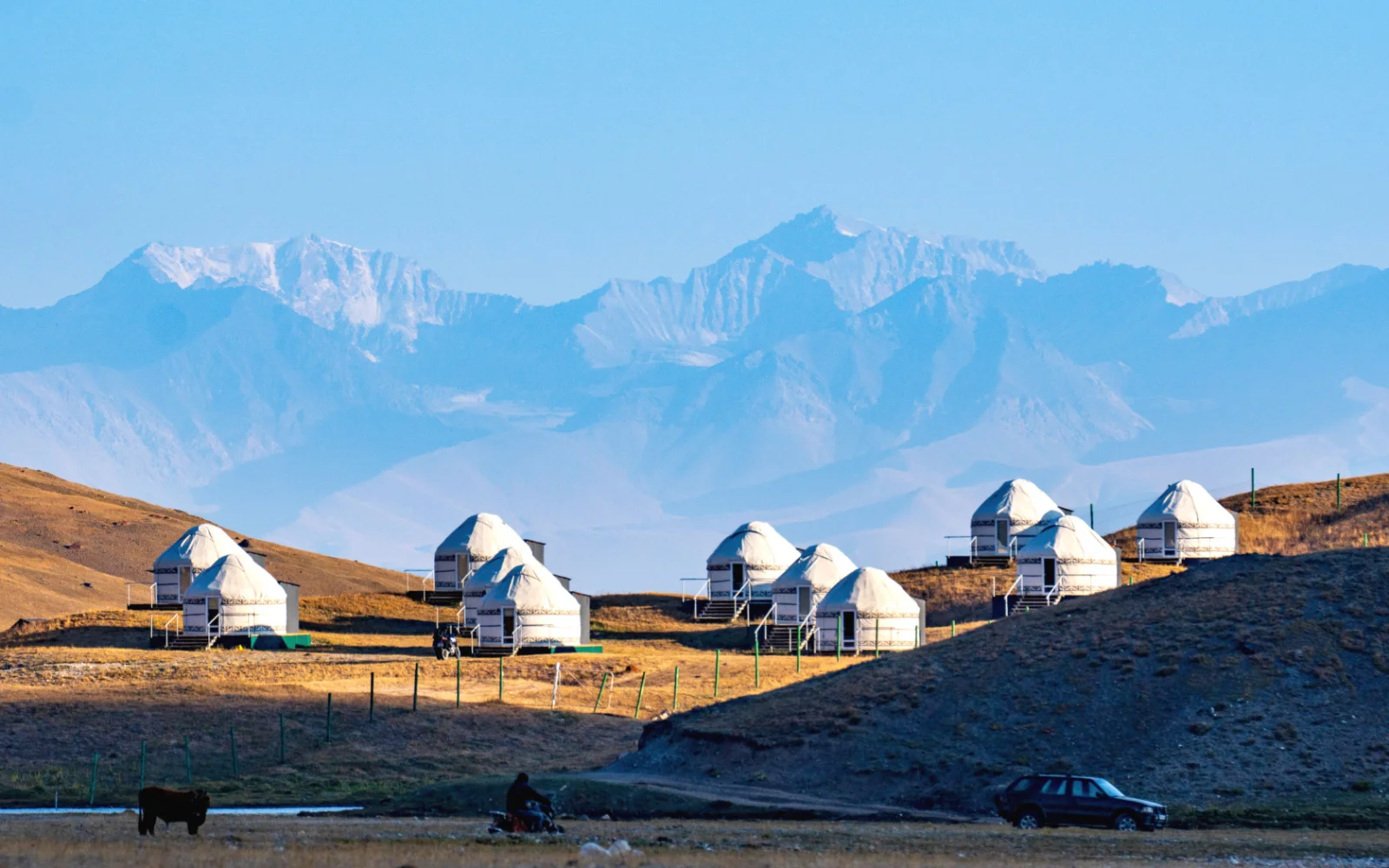 Jurtencamp in der Hochgebirgswelt des Pamir in Kirgisistan mit weißen Jurten im Licht der aufgehenden Sonne auf einer kargen Hochweide vor schneebedeckten Bergen