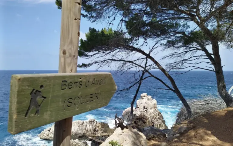 Holzwegweiser nach Bens d'Avall und Sóller an einem Küstenpfad mit Blick auf das blau-weiße Meer, Felsen und Pinien.