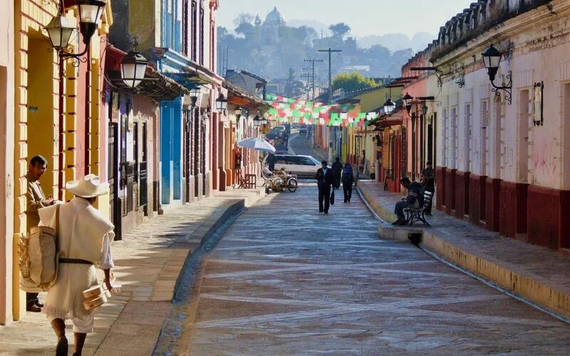 Gepflasterte Kolonialstraße in in San Cristóbal de las Casas mit bunt angestrichenen Häuserfassaden, Straßenlaternen und einigen Fußgängern, darunter ein Mann in traditioneller weißer Kleidung und Hut links im Vordergrund.